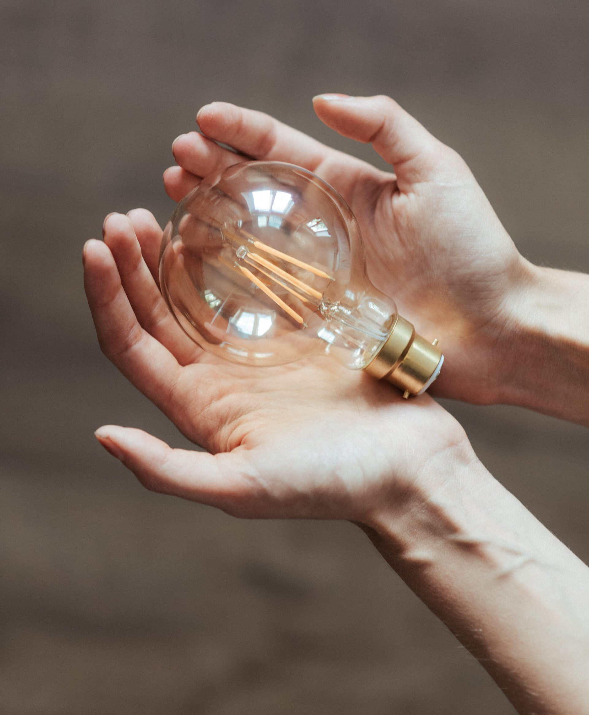 Hands cupped around a clear lightbulb, held against a brown wooden background.