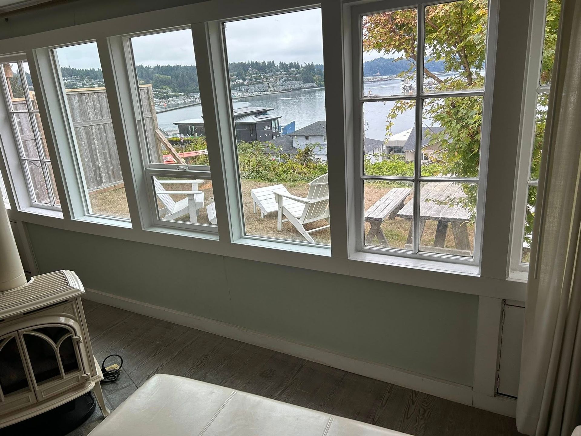 Living room with large windows overlooking a harbor.  White trim, light green walls, two white chairs, and a picnic table are visible.