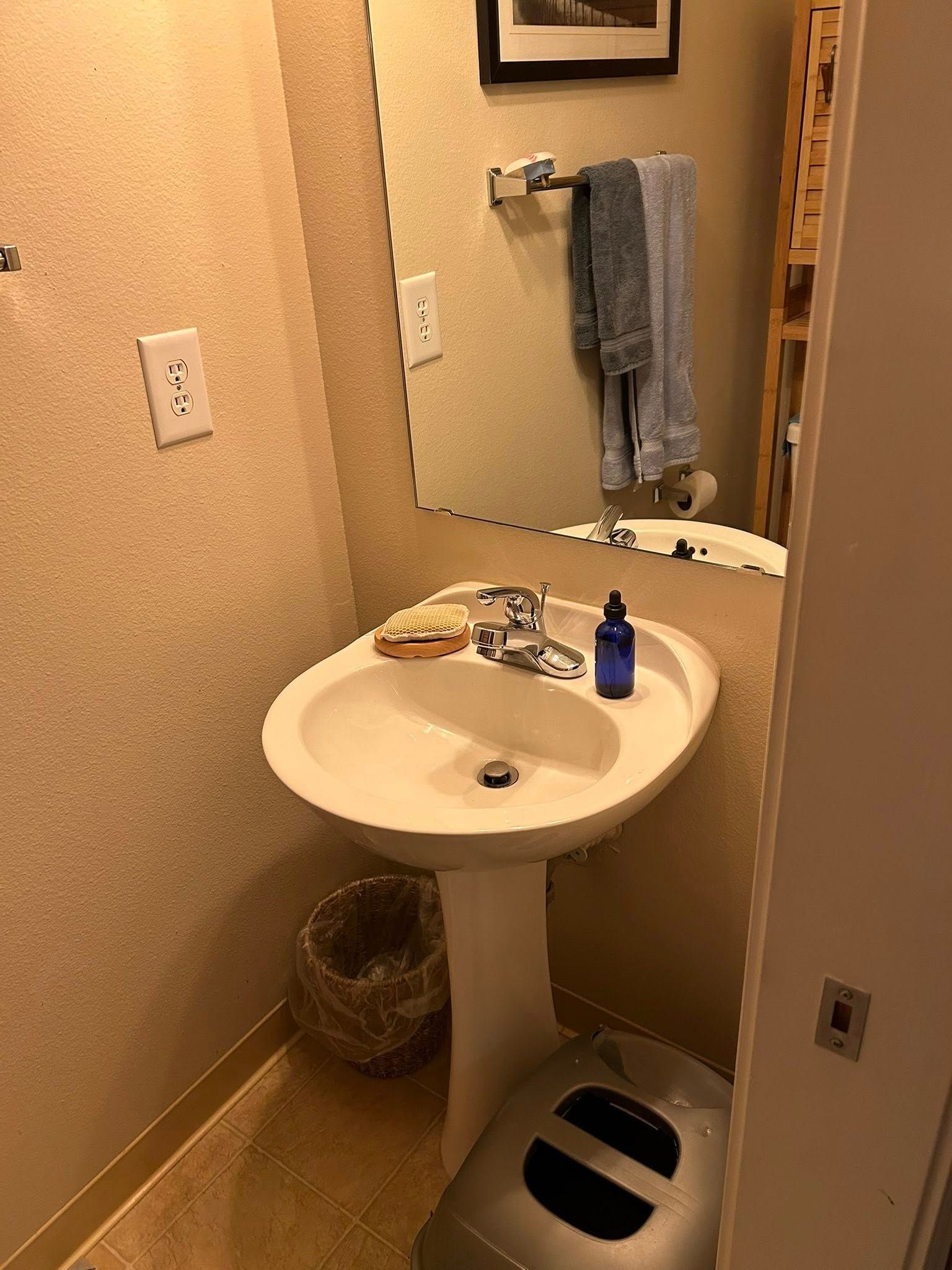 Bathroom with pedestal sink, mirror, towels, and a trash can. Light tan walls and floor.