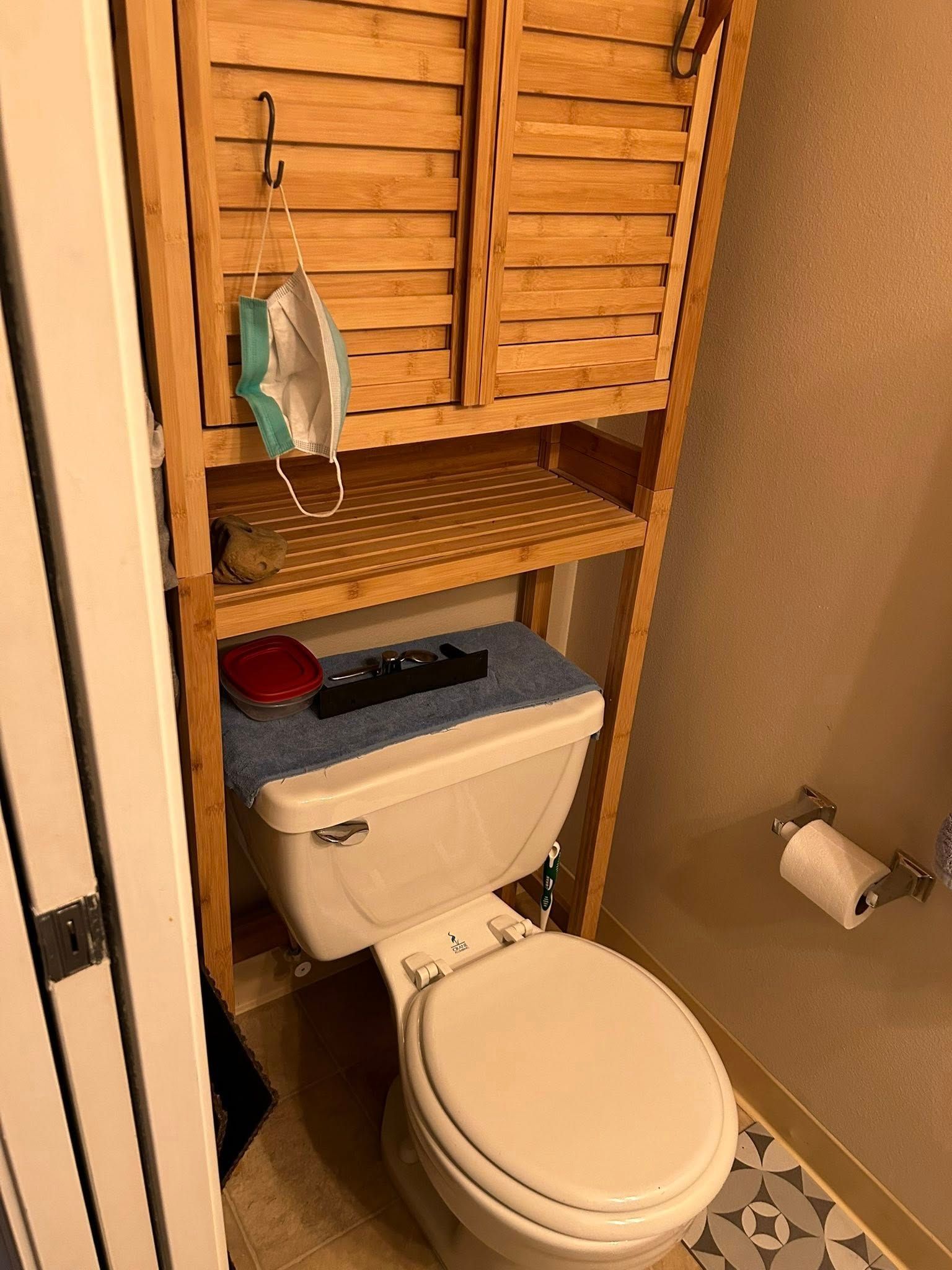 Wooden cabinet over a toilet. A mask hangs on a hook. A remote sits on the tank.