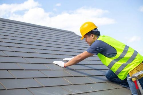 Roofer wearing a yellow helmet and safety vest inspecting dark gray roof tiles on a sunny day.