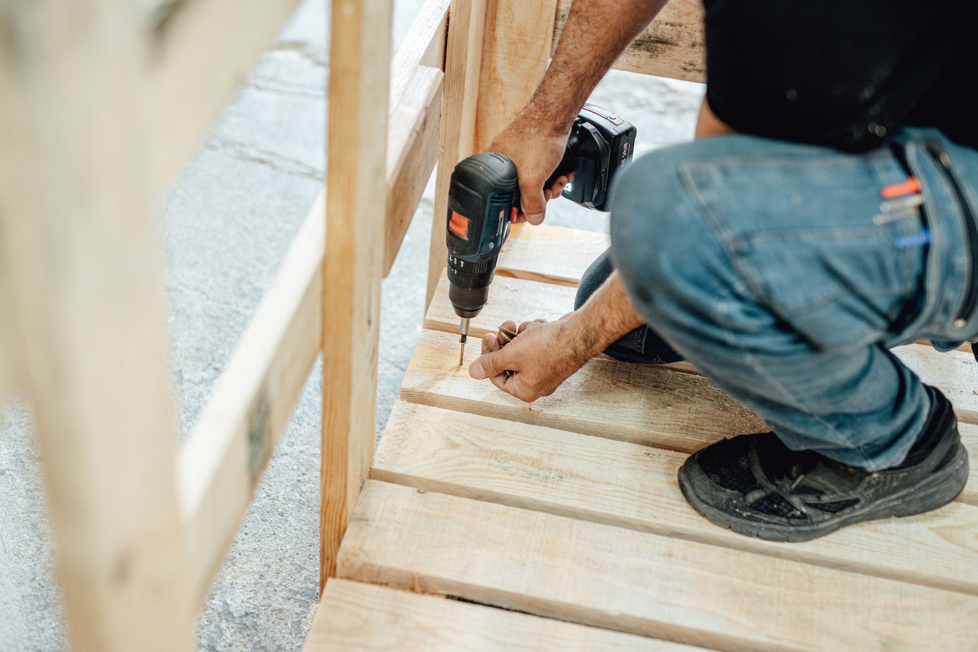Person in jeans and black shirt using a drill to screw wood planks together outdoors.