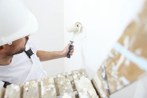 Painter in white overalls and hard hat painting a wall with a roller.