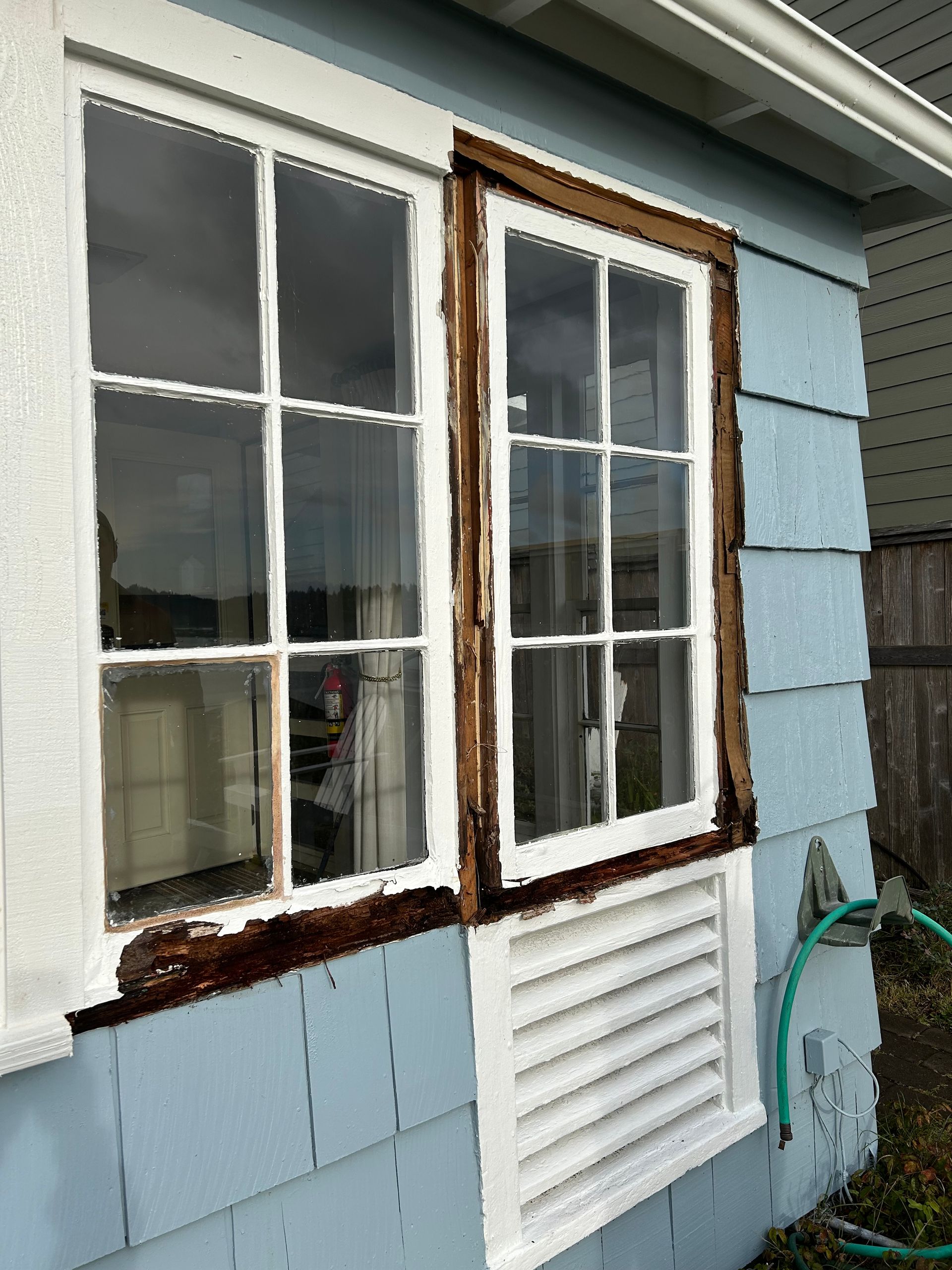 Exterior view of a window with rotted wood trim on a light blue building.