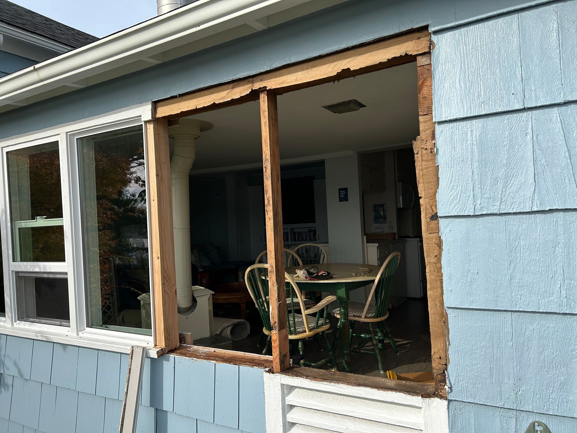 Window opening in blue siding, wooden frame exposed. Interior with table and chairs visible.