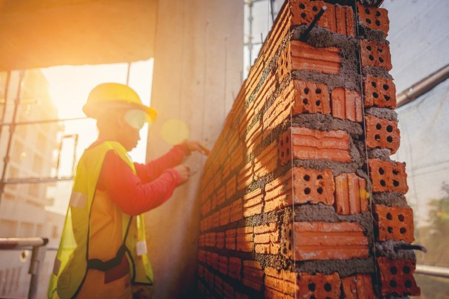 Construction worker laying bricks on a wall at sunrise, wearing a hard hat and reflective vest.