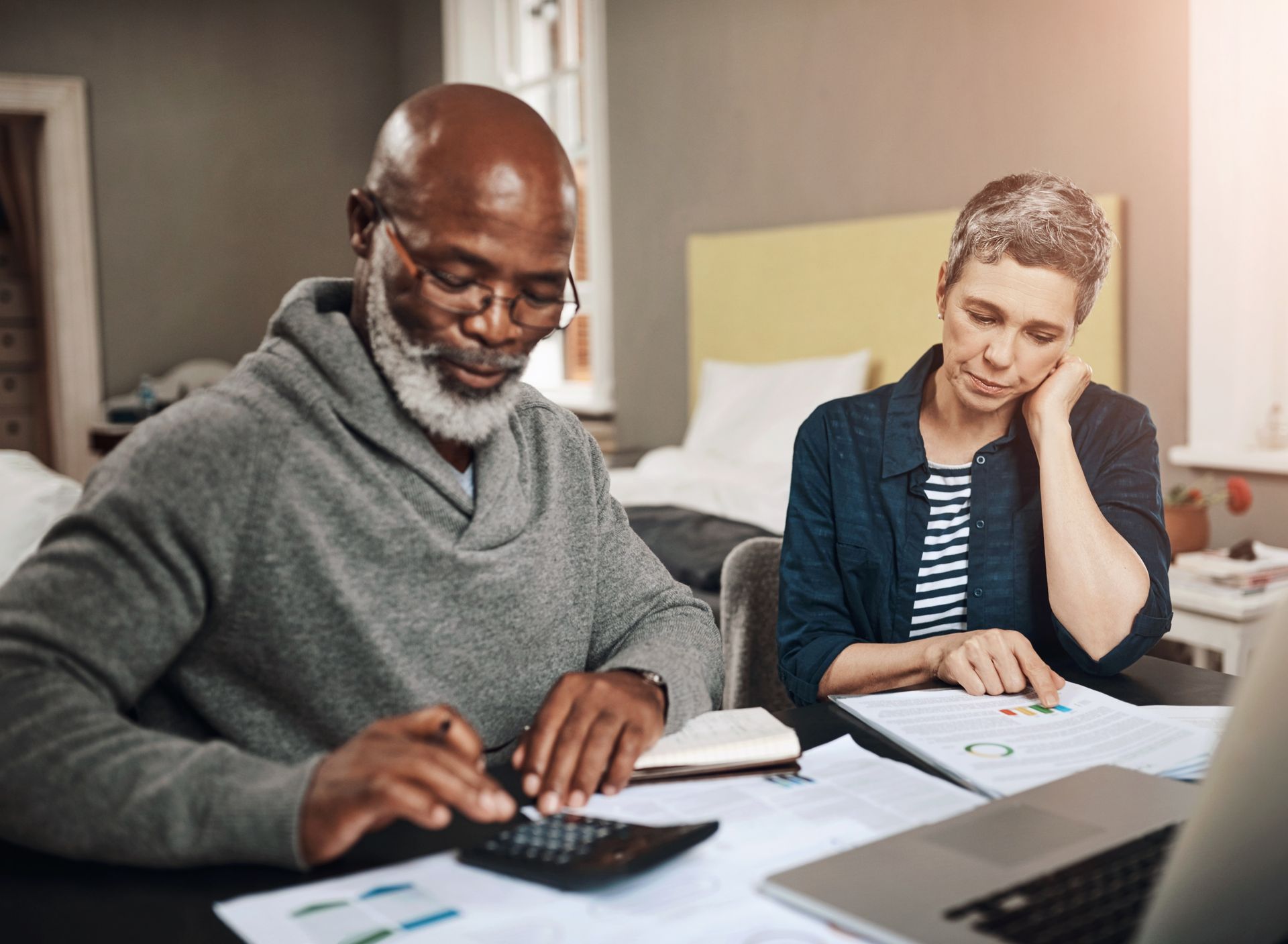Man using a calculator, woman points to documents. Both appear to be focused, reviewing finances.