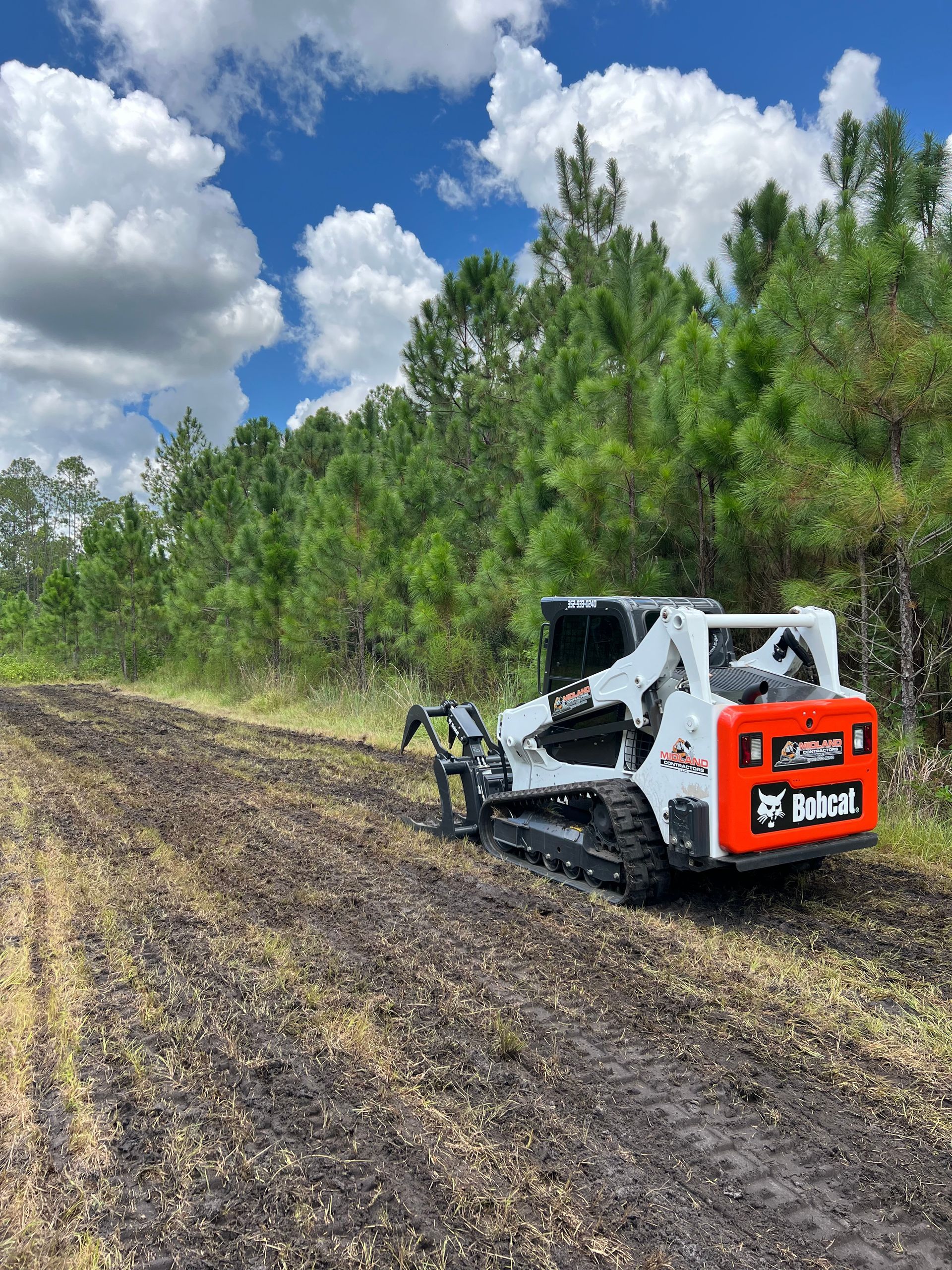 A bobcat is plowing a field with trees in the background. - Midland Contractors - Clermont, FL