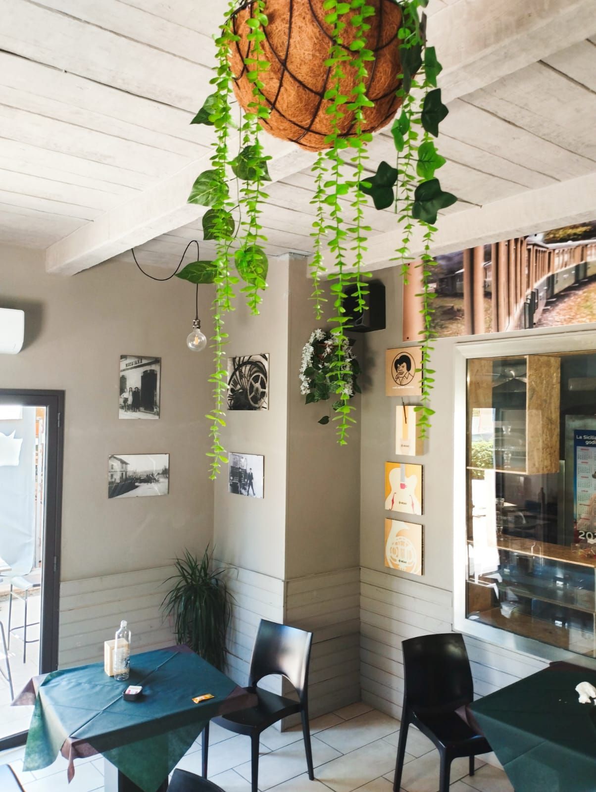 Interno del bar con un cesto di piante appeso, sedie nere e quadri incorniciati alle pareti. I tavoli sono rivestiti di tovaglie verdi.