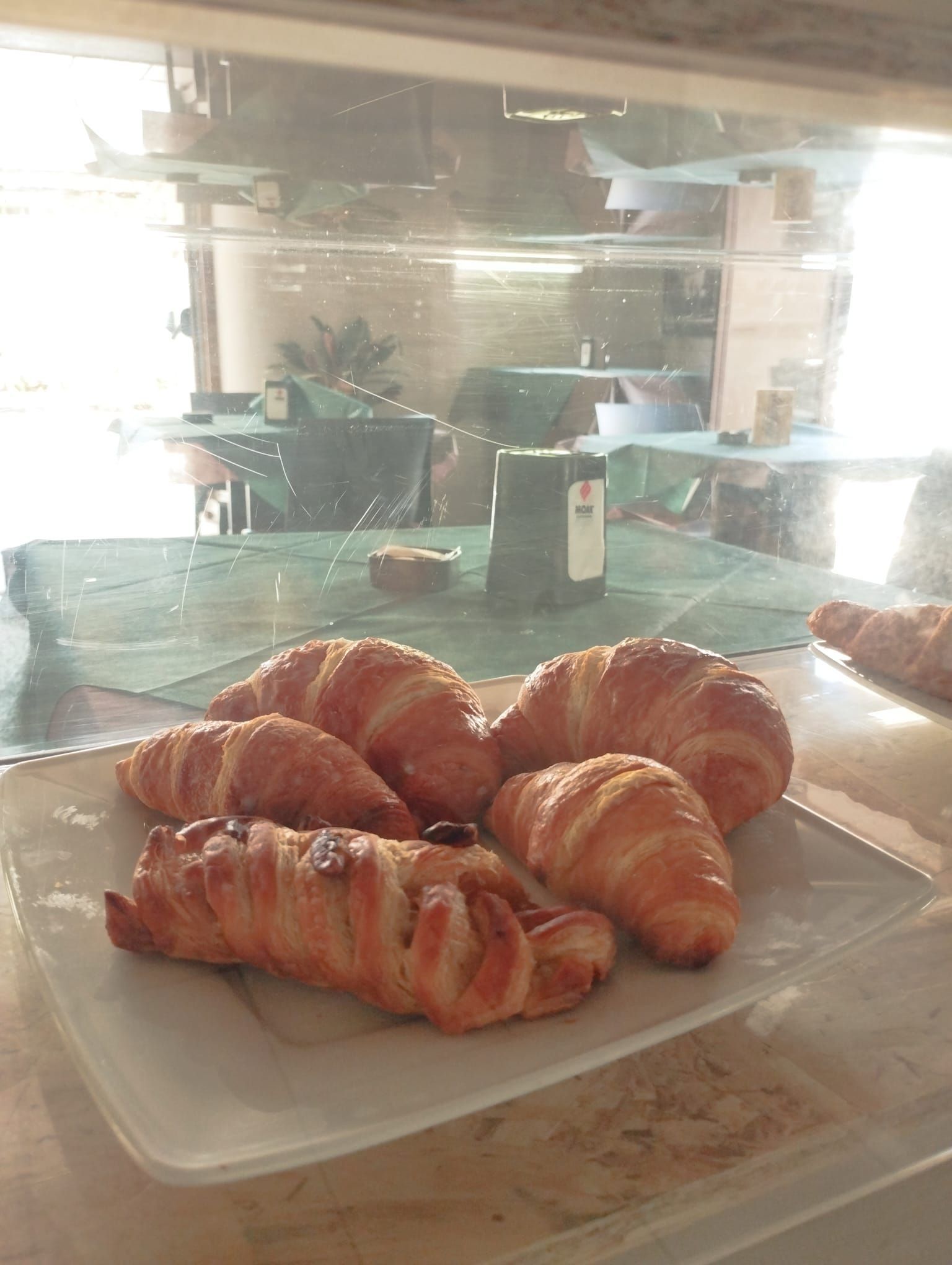 Dietro un vetro si trovano un piatto di croissant e un dolce, che riflette l'immagine di un bar con tavolini verdi.
