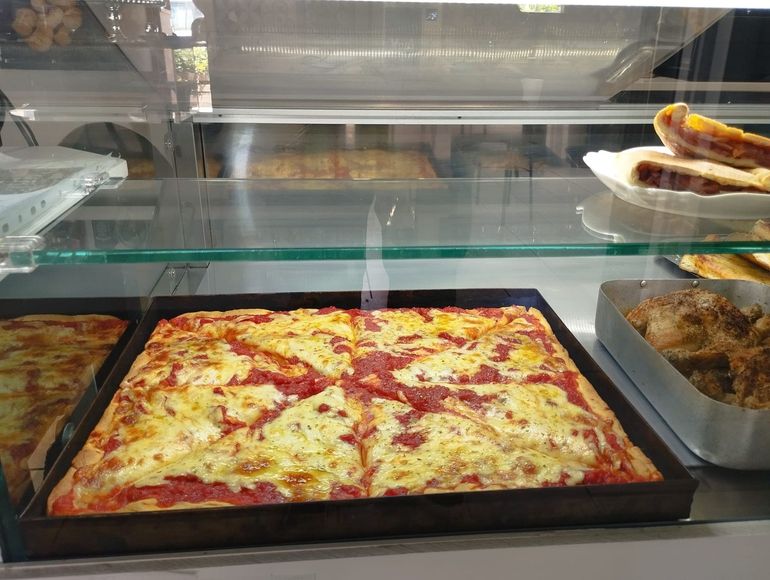 Una pizza quadrata con formaggio fuso e salsa di pomodoro è esposta in una vetrina, insieme ad altri prodotti da forno.