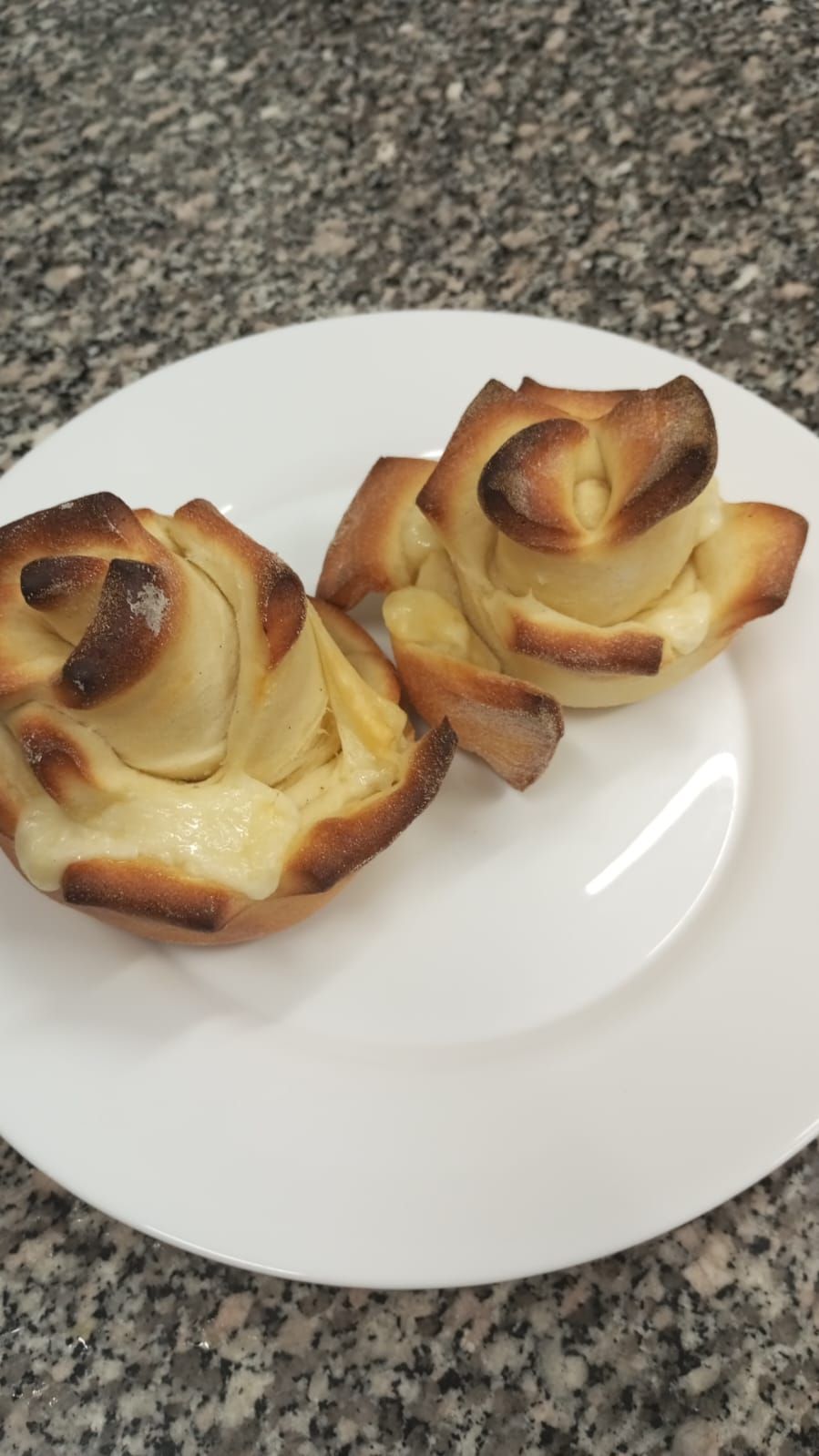 Due rose di pasta sfoglia cotta al forno su un piatto bianco, appoggiate su un piano di lavoro screziato. Le rose sono di un marrone dorato con strati visibili.