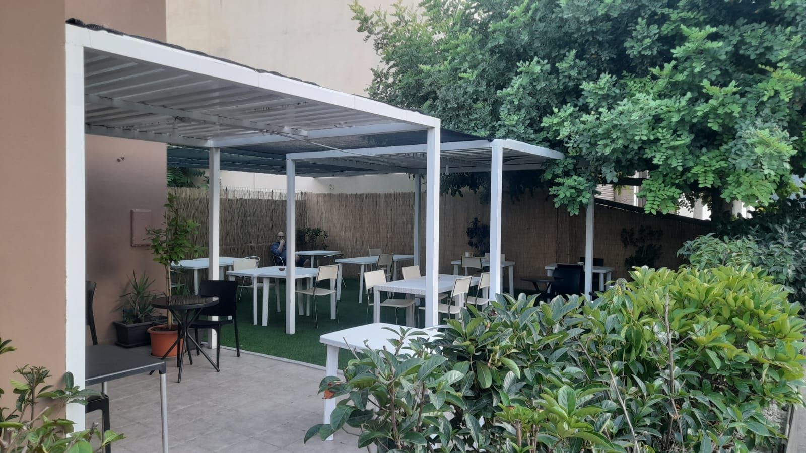 Area esterna con pergolati bianchi, tavoli e sedie. Arbusti e alberi verdi circondano lo spazio.