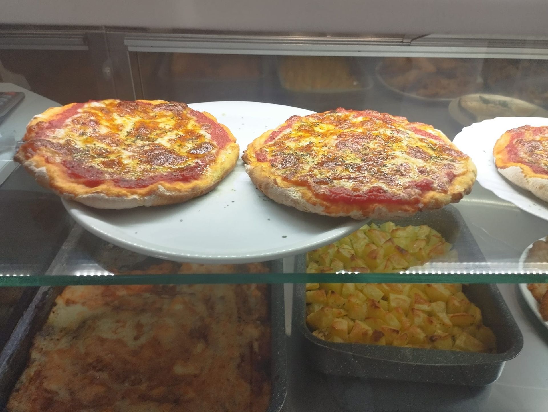 Due piccole pizze su un piatto in una vetrina, insieme ad altri alimenti come patate e quella che sembra essere lasagna.