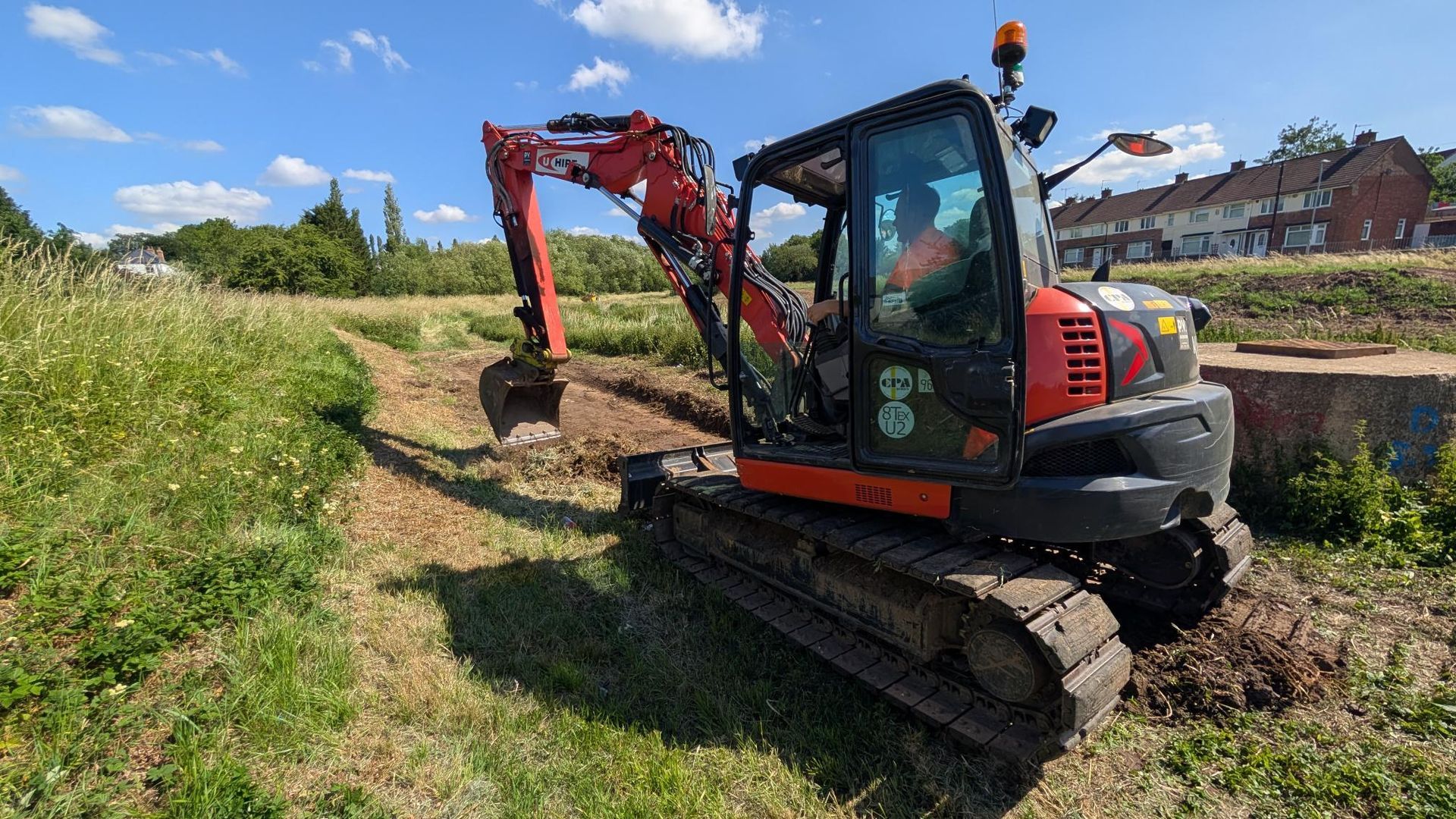 A red compact excavator digs a trench in a grassy field under a sunny blue sky, with houses visible in the background.