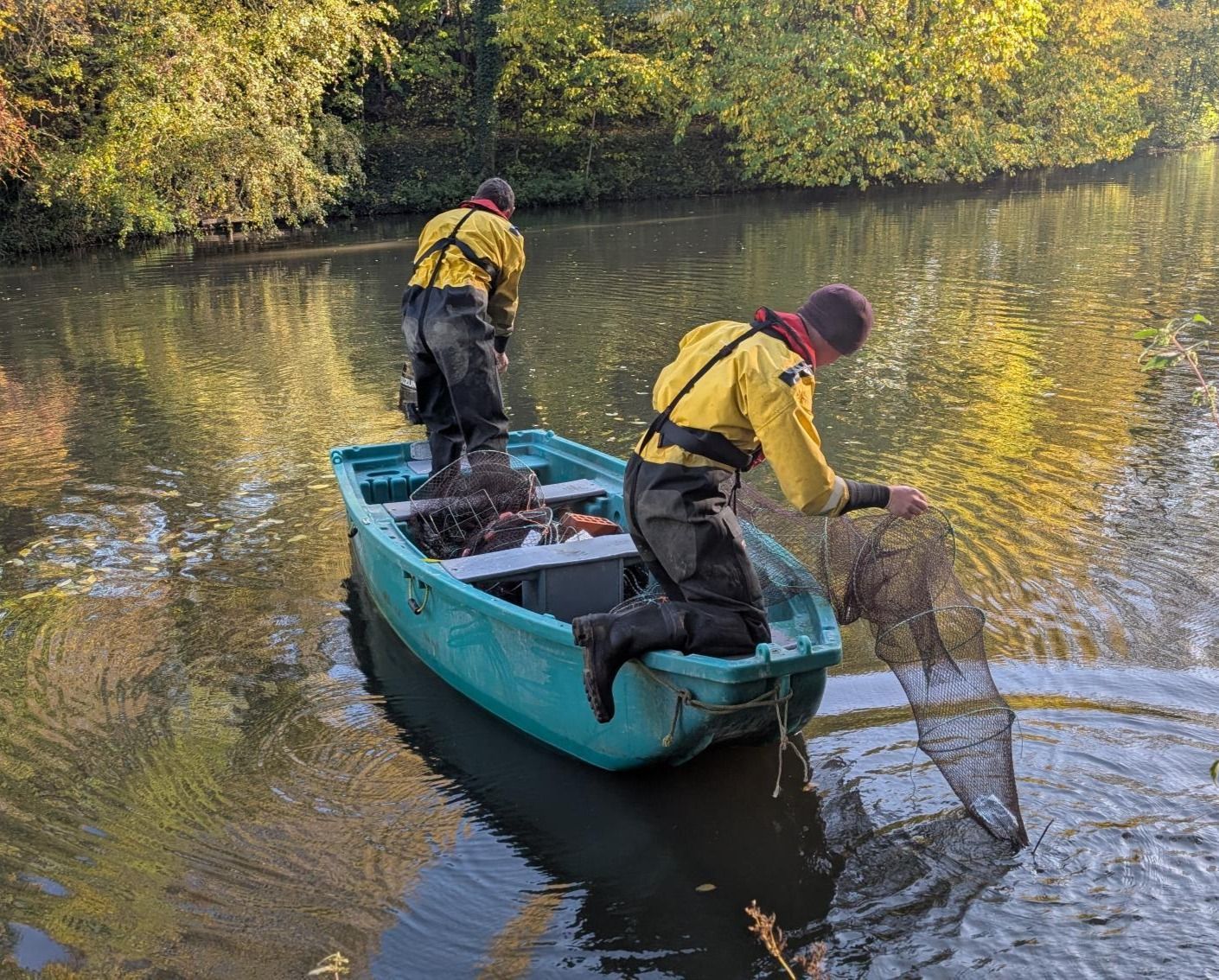 Two people in yellow waterproof gear and waders pull a large net from a boat on a calm river surrounded by autumn trees.