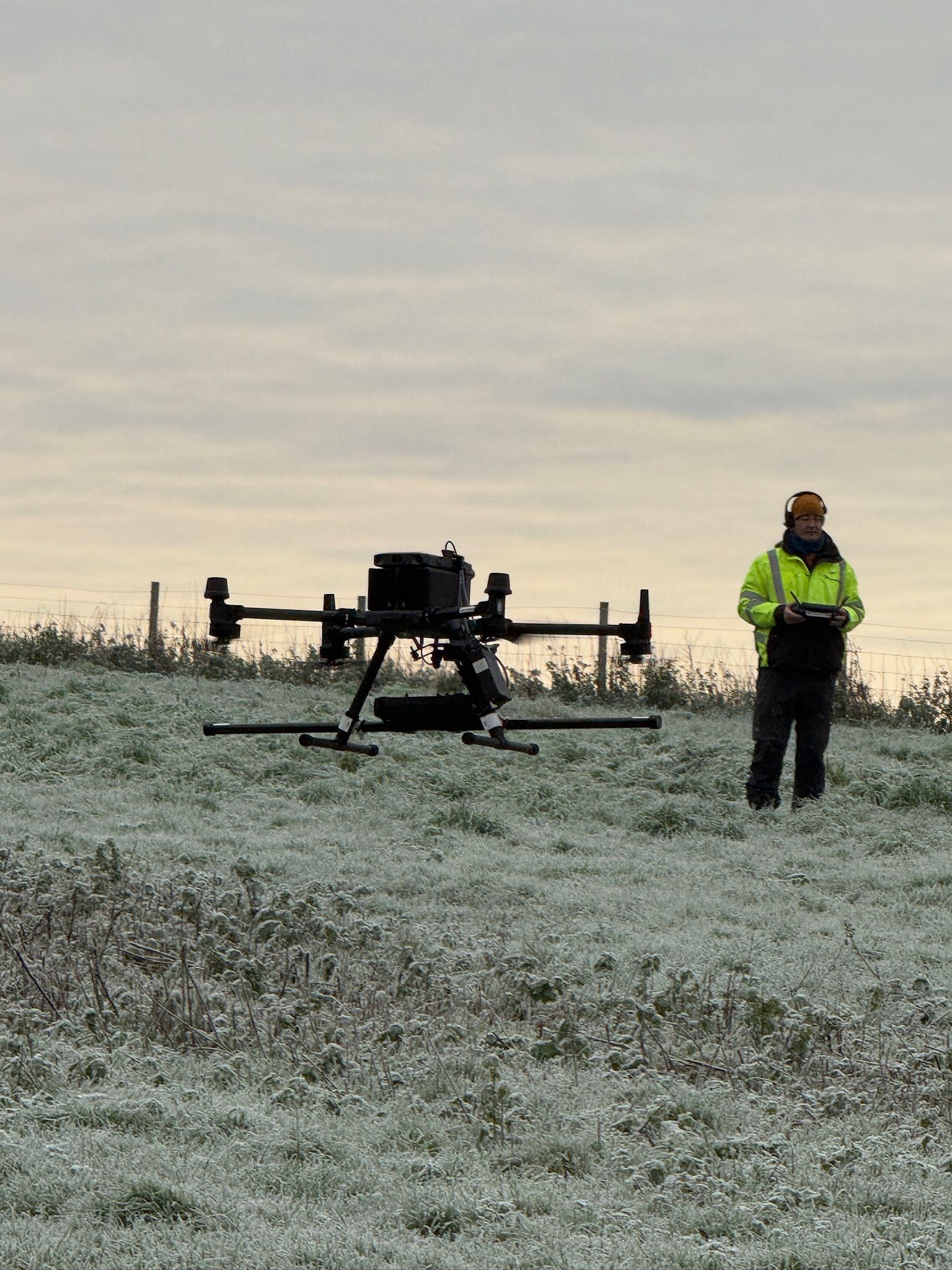 A person in a high-visibility jacket operates a professional drone in a frost-covered, grassy field at dawn.