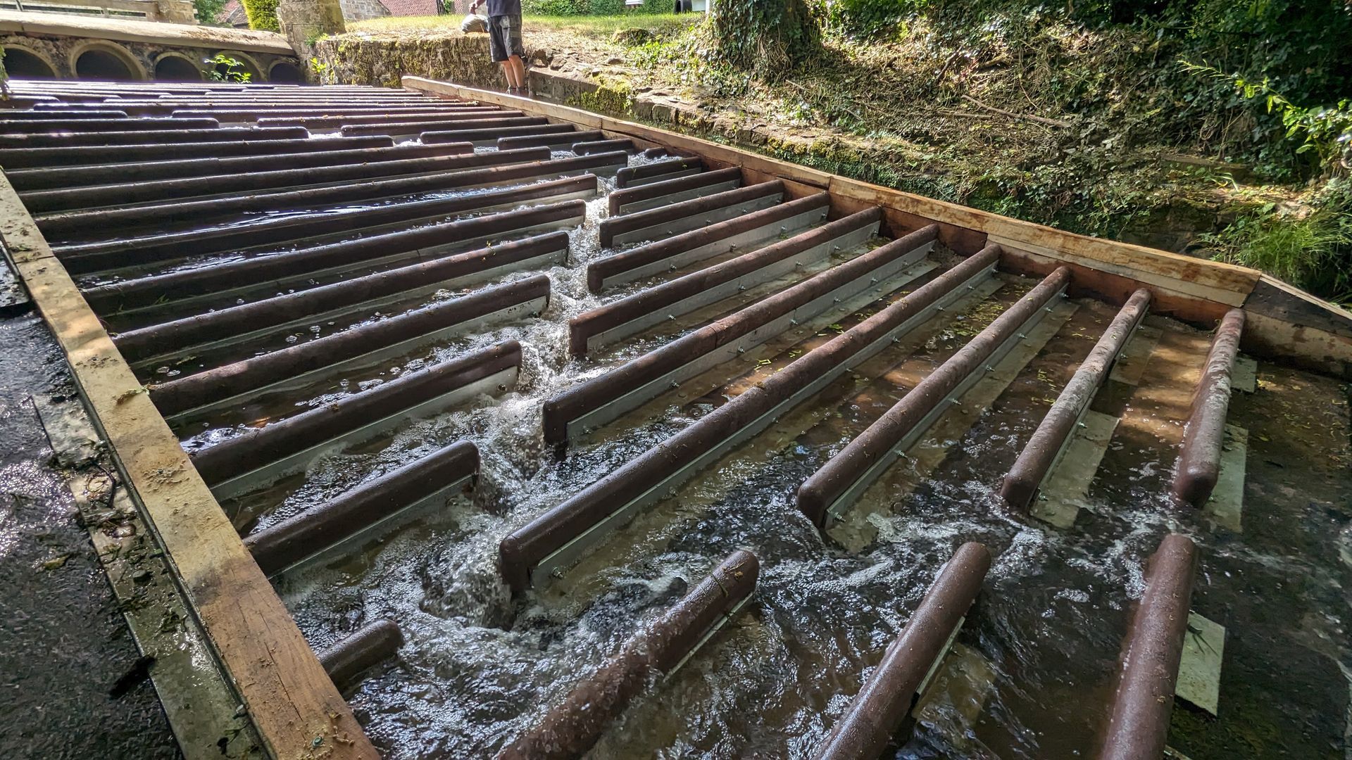 Water flows down a tiered concrete spillway, cascading over metal baffles set in a zigzag pattern across the slope.