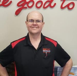 A bald man wearing glasses and a black and red shirt is standing in front of a sign that says `` we good to ''.