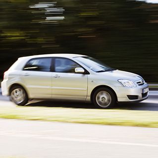 A silver car is driving down a road with trees in the background