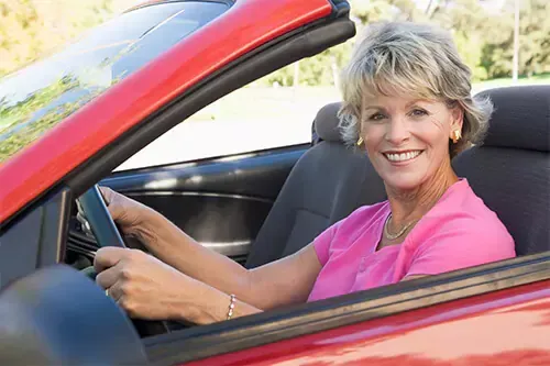 A woman in a pink shirt is driving a red car and smiling.