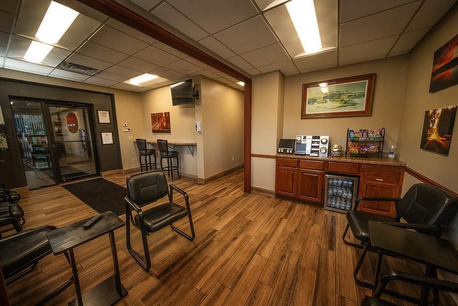 A waiting room with wooden floors , chairs , tables and a refrigerator.