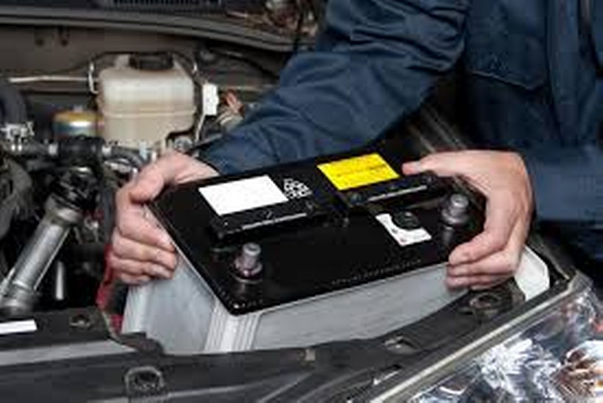 A man is holding a car battery under the hood of a car.