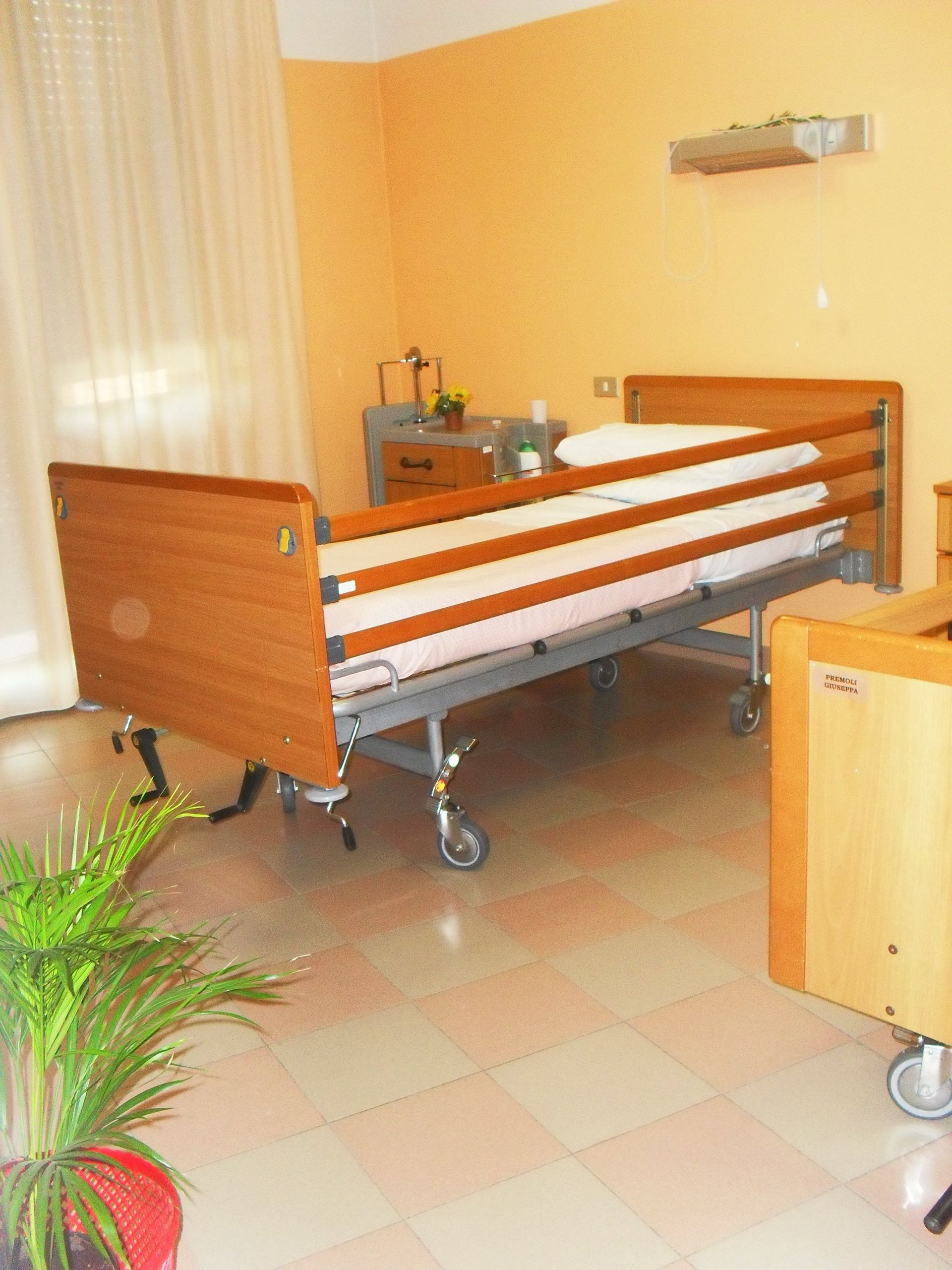 Stanza d'ospedale con letto regolabile, testiera in legno, pareti beige e pavimento piastrellato.