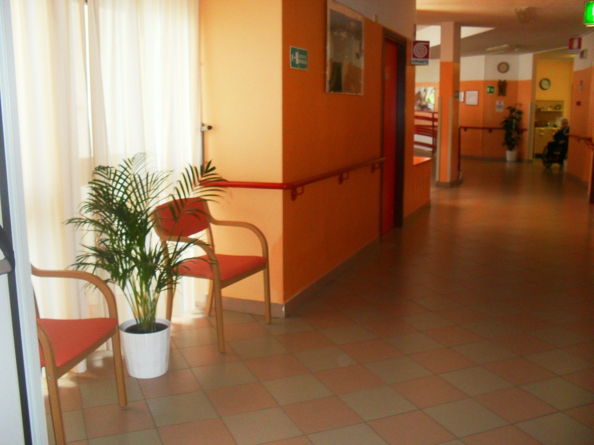 Corridoio interno con pareti arancioni, sedie, una pianta in vaso e un lungo corridoio che conduce alle stanze.
