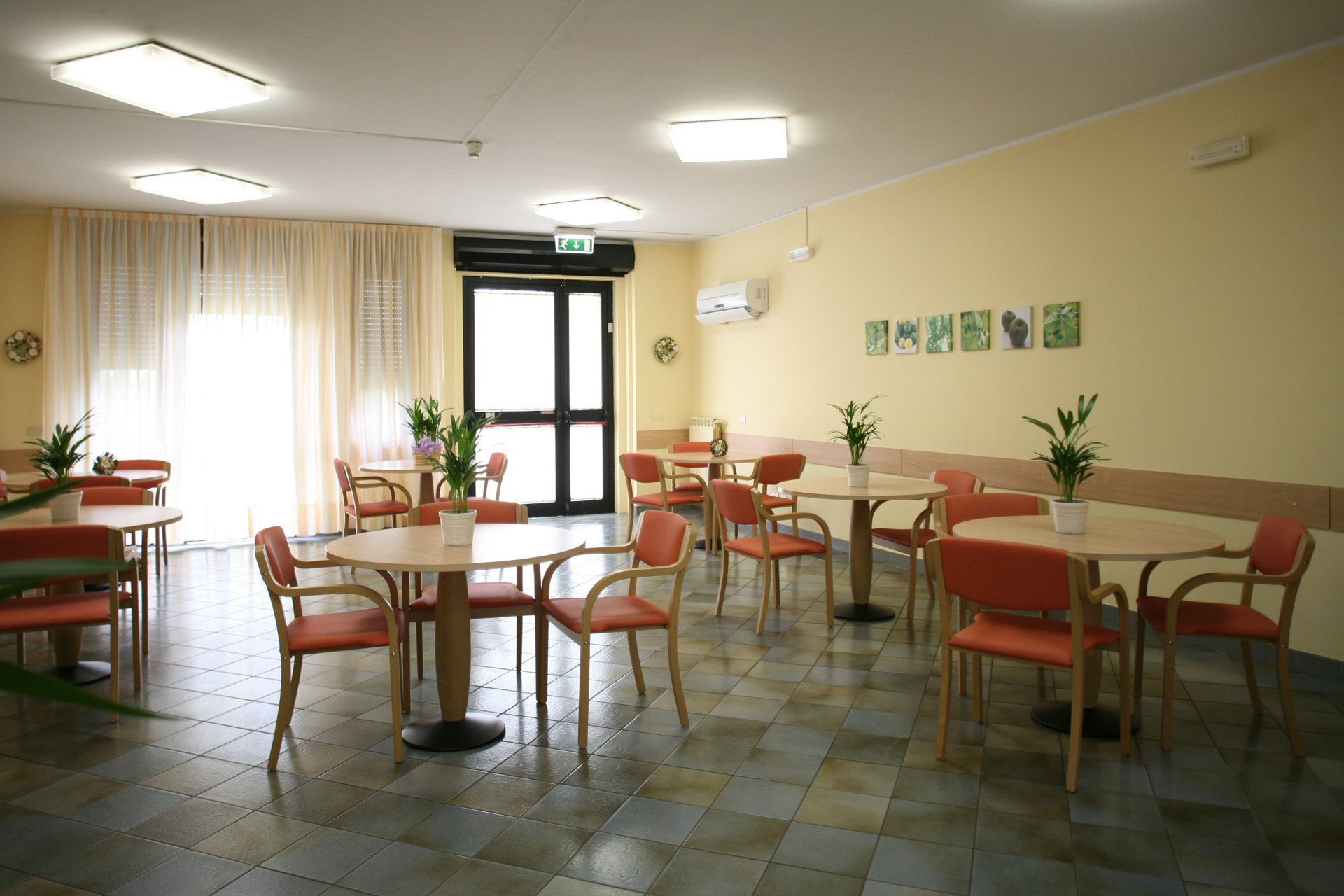 Zona salotto in stile bar: tavoli rotondi, sedie arancioni, pavimento piastrellato, pareti chiare e tende trasparenti.