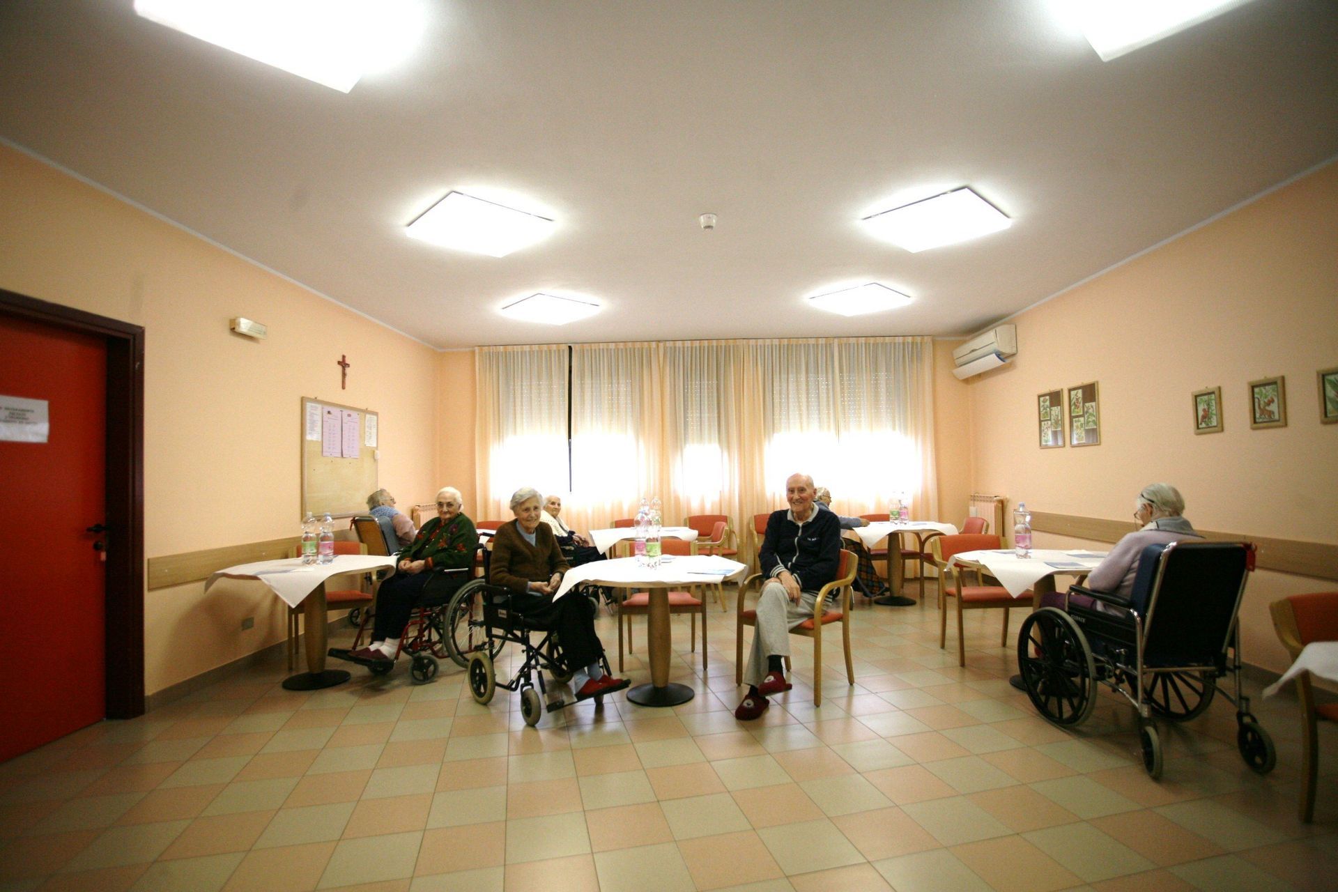 Anziani su sedia a rotelle e sedie seduti ai tavoli in una sala da pranzo ben illuminata, con tende e una porta rossa.