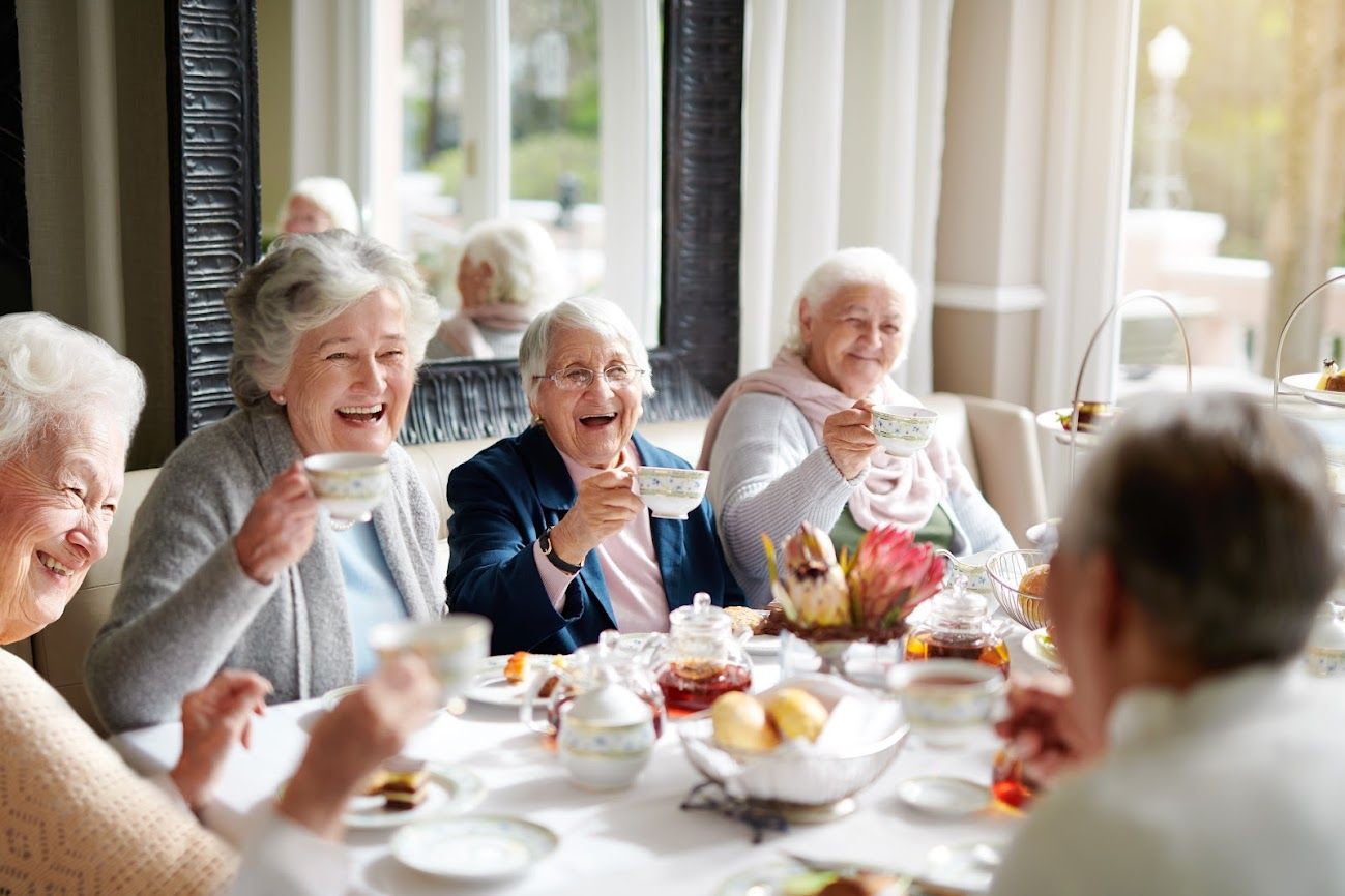 Donne anziane che ridono, sorseggiando il tè sedute a un tavolo in una luminosa sala da pranzo.