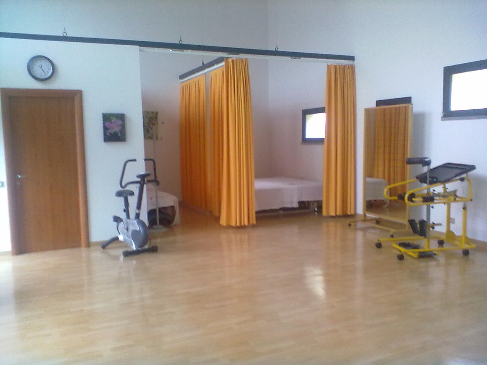 Sala di fisioterapia con attrezzi ginnici, letto dietro tende gialle e orologio.