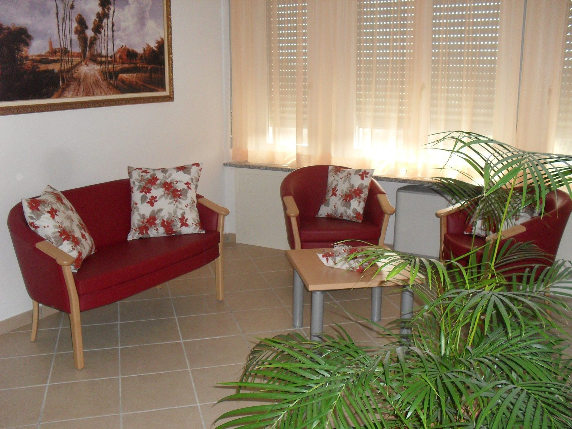 Zona salotto rossa con cuscini floreali, tavolino e verde. Quadro e finestra con tende.