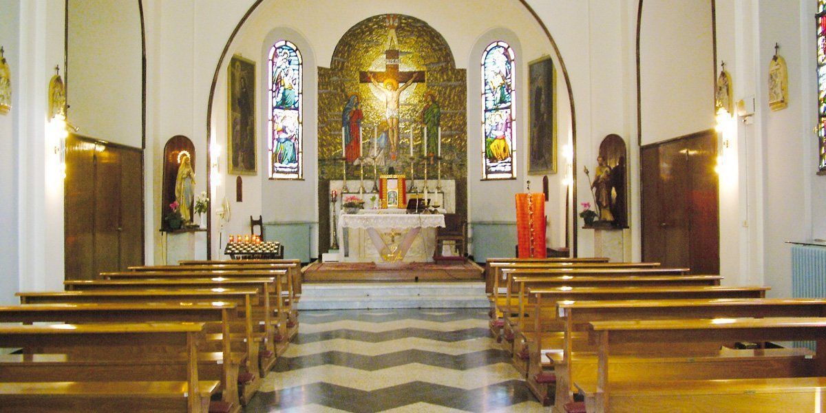 Vista interna di una chiesa con file di banchi rivolti verso l'altare con un crocifisso e vetrate colorate.