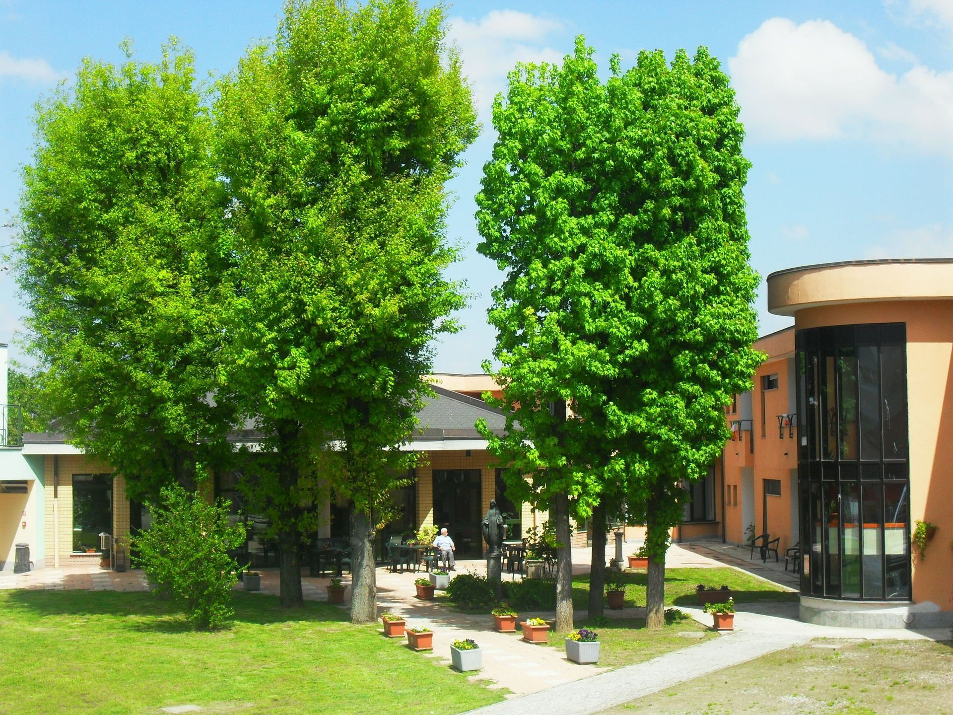 Lussureggianti alberi verdi di fronte a un edificio, con persone sedute all'esterno, in una giornata di sole.