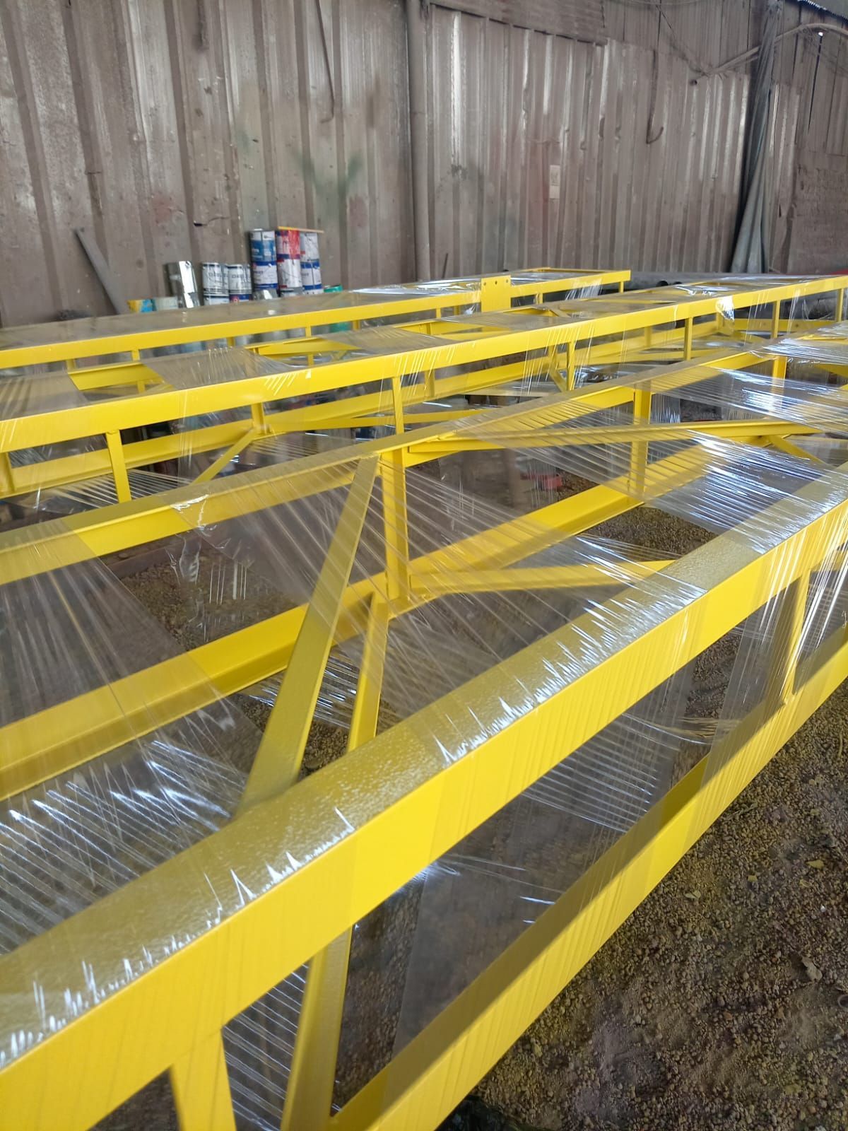 Marcos de metal amarillo, posiblemente andamios o estanterías, con envoltura de plástico, dentro de un taller.