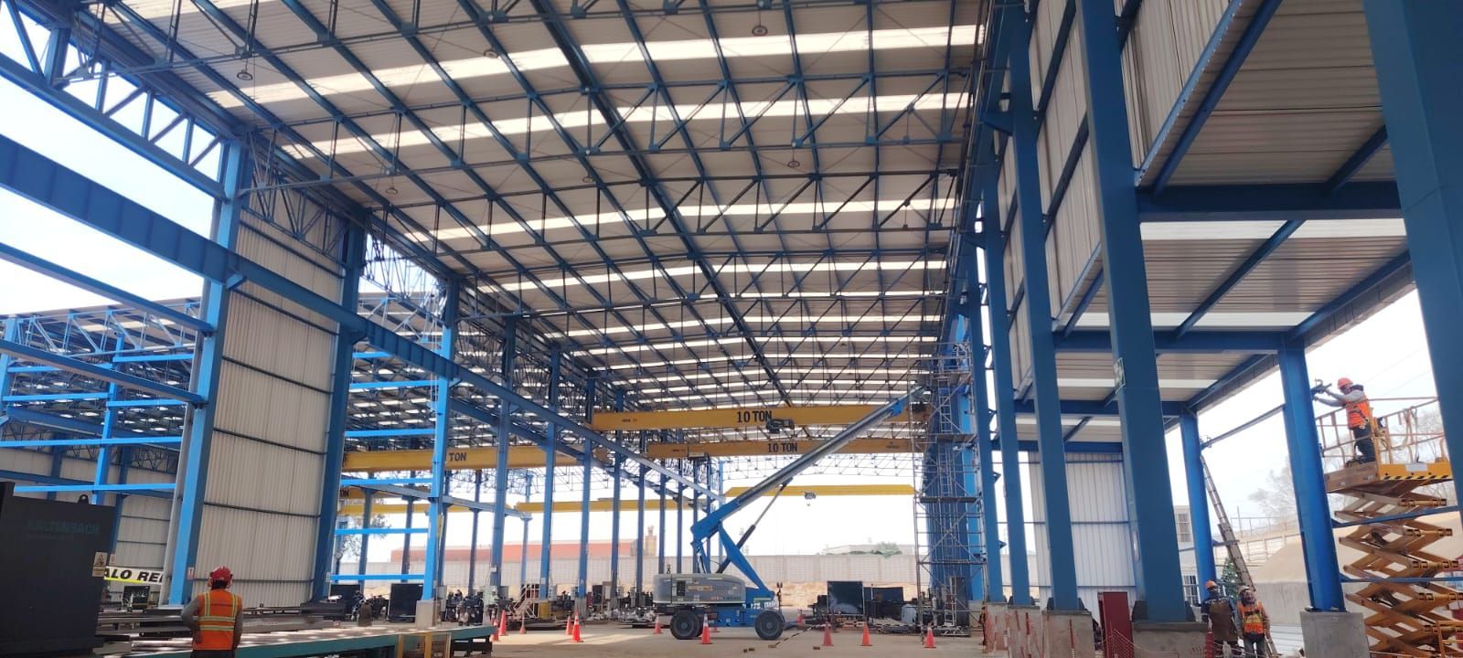 Gran edificio industrial en construcción; soportes de acero azul; trabajadores, ascensor y grúa aérea presentes.