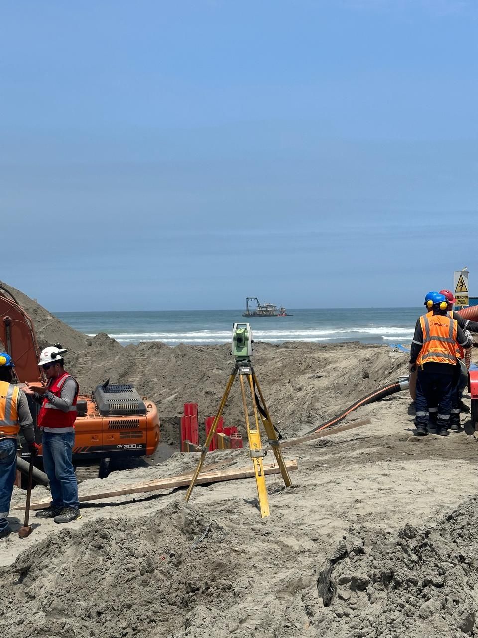 Topógrafos en un sitio de construcción cerca del océano, utilizando equipos y trabajando en suelo arenoso.