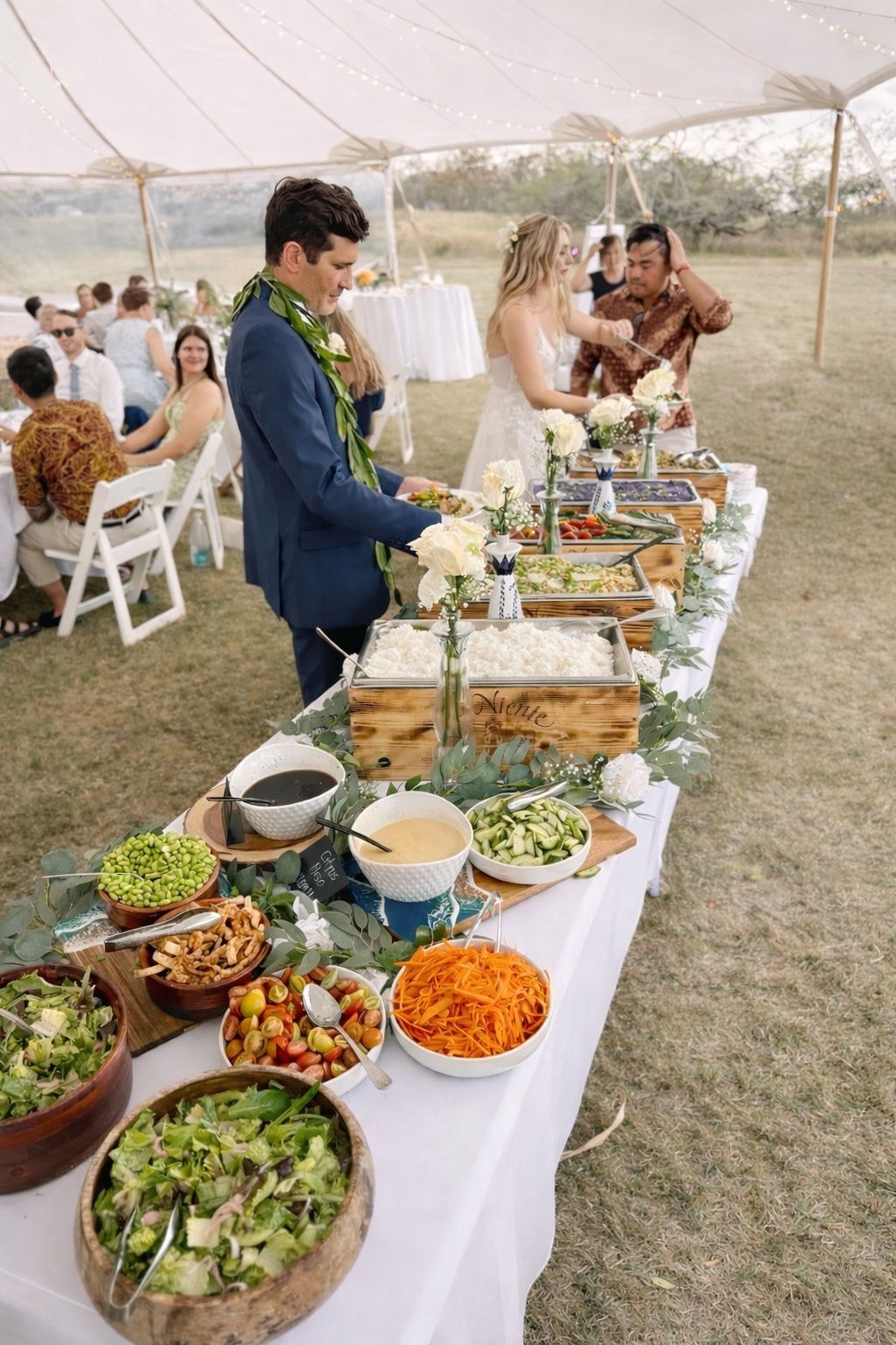 Outdoor buffet table at a wedding reception, with guests serving food under a tent.
