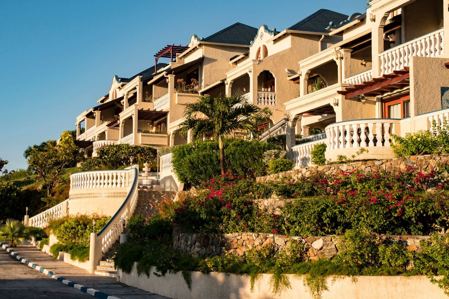 Terraced beige buildings with balconies and white railings, surrounded by lush greenery and flowers under a blue sky.