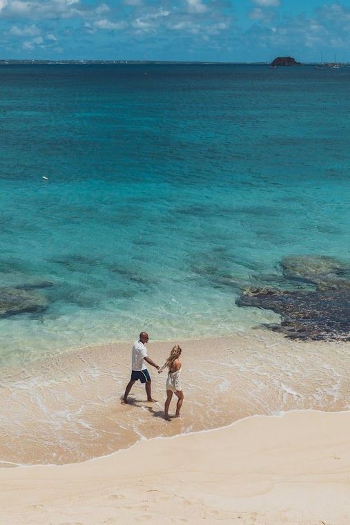 Couple walking hand-in-hand on a beach, turquoise water, bright blue sky, small island in distance.