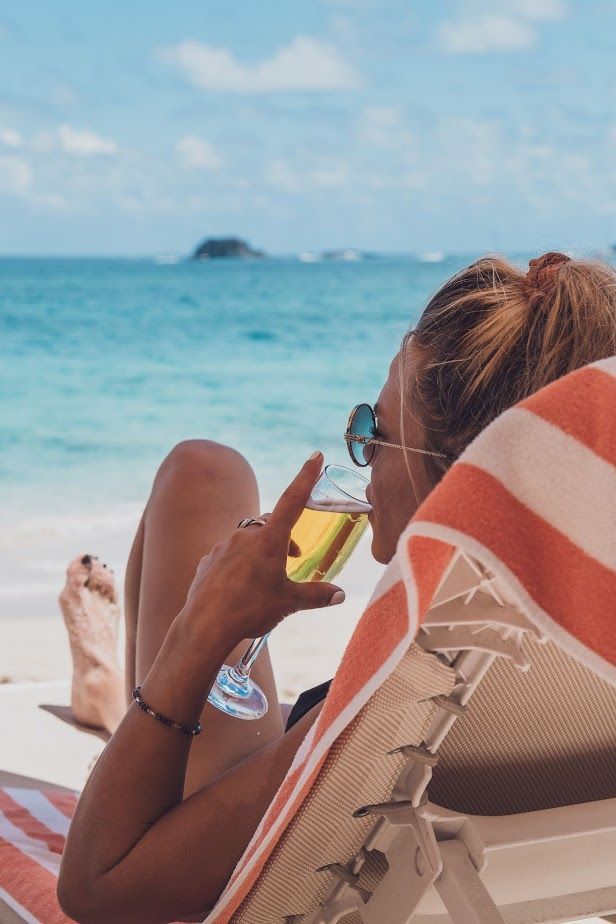 Woman in sunglasses sips drink while relaxing on a beach chair, ocean in the background.