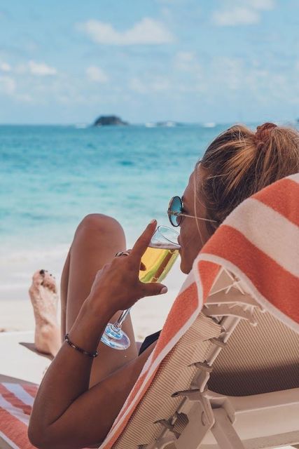 Woman in sunglasses sips drink while relaxing on a beach chair, ocean in the background.