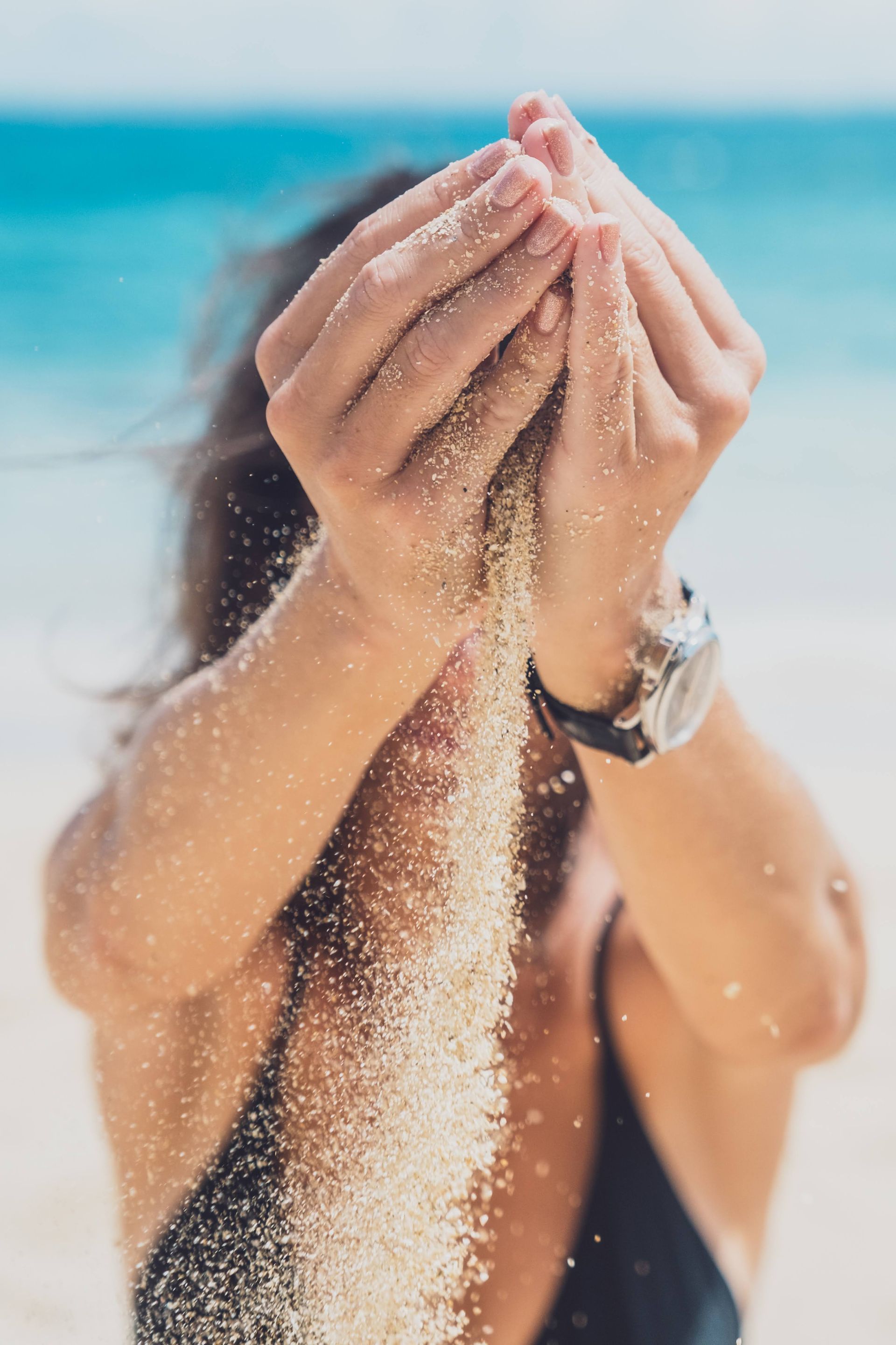Woman on beach, holding up hands as sand pours through, blue water in background.