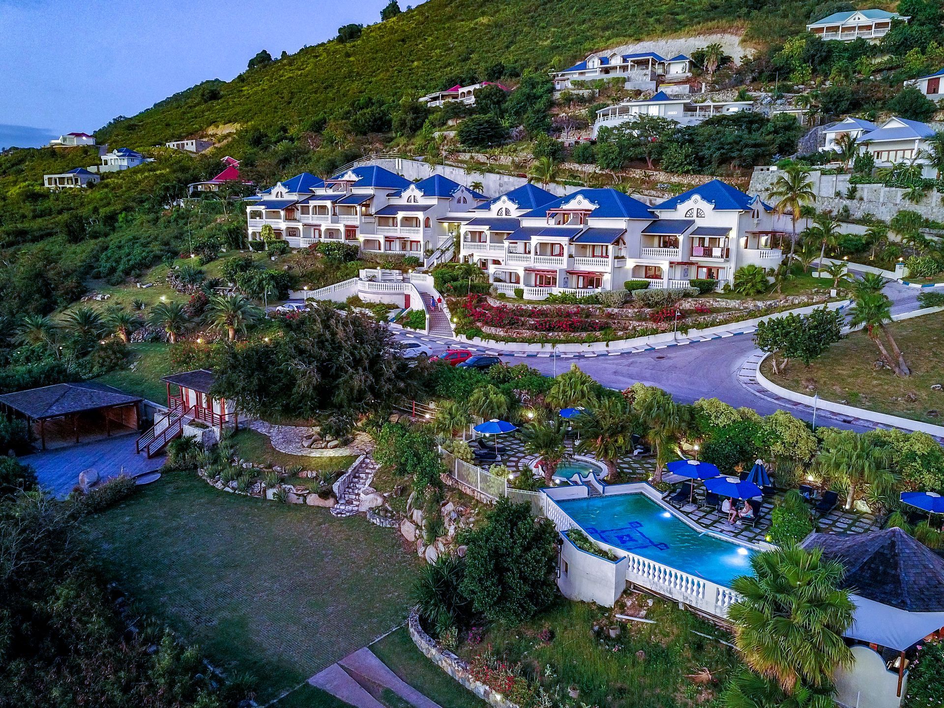 Luxury resort nestled on a hillside with white buildings, blue roofs, pool, and lush greenery.
