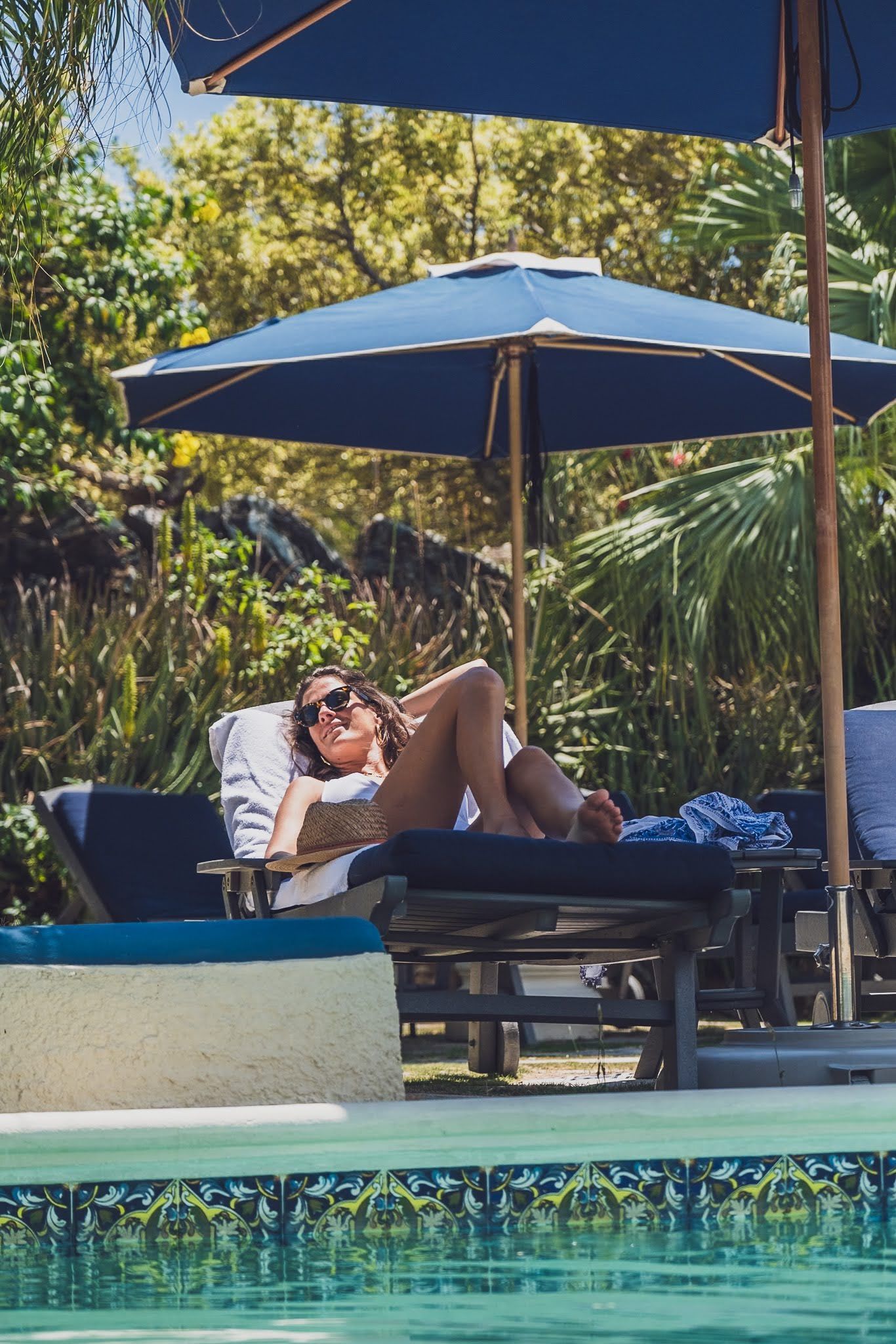 Woman sunbathing on a lounge chair by a pool, under a blue umbrella.
