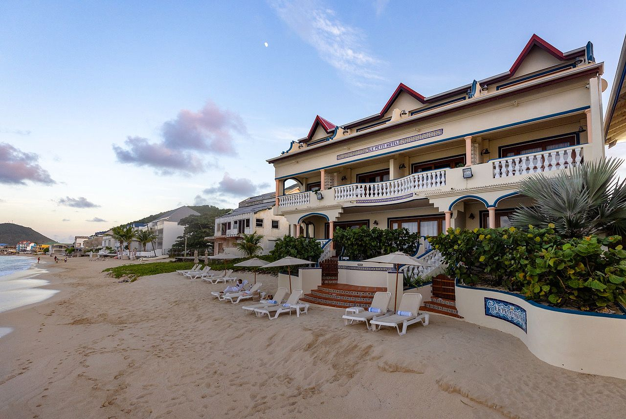 Beachfront hotel with beige exterior, blue accents, lounge chairs, and ocean view under a partly cloudy sky.