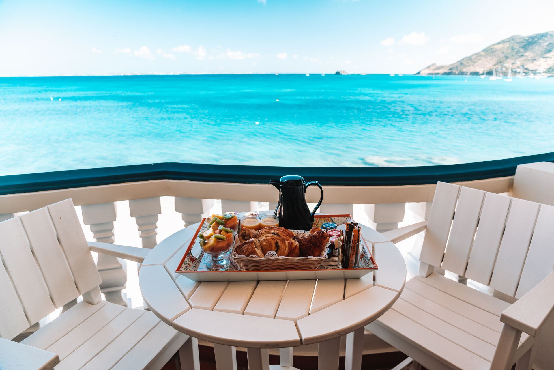 Breakfast tray on a white table, overlooking a turquoise ocean from a balcony.