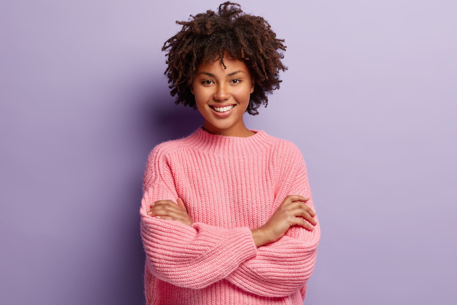 A woman in a pink sweater is standing with her arms crossed and smiling.